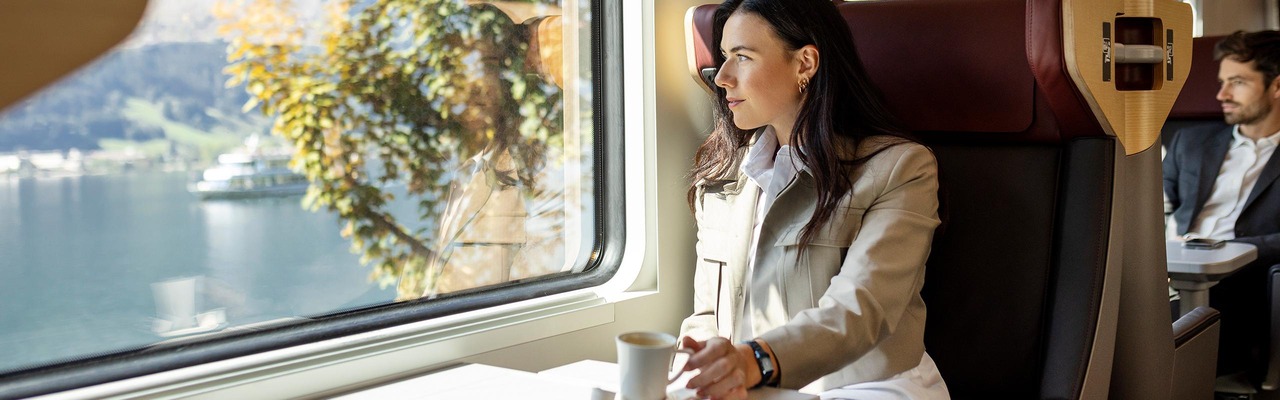 Frau sitzt mit Tasse Kaffee im Railjet und blickt aus dem Fenster