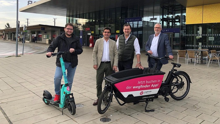 Personen präsentieren Lastenrad und E-Scooter vor Bahnhofsgebäude