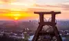 Vor dem Hintergrund der aufgehenden Sonne in Essen erhebt sich der Doppelbock-Förderturm der Zeche Zollverein in Essen. Darunter erstrecken sich Häuserzeilen und grüne Bäume. Der Himmel ist orange und gelb gefärbt.<br/>