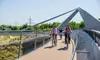 Mehrere Personen fahren mit Fahrrädern über eine moderne Fuß‑ und Radbrücke mit geschwungenen Metallstreben; seitlich verlaufen Geländer, im Hintergrund sind Gleise, Grünflächen und Strommasten unter klarem Himmel zu sehen.<br/>