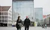 Zwei Personen gehen über einen Platz vor einem modernen Glasgebäude in der Innenstadt, Familie mit Kinderwagen im Hintergrund.<br/>