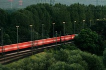 Red high-speed train passing through green forest landscape with overhead power lines.