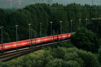 Passing container train with red containers blurred by speed, set against a green forest backdrop.