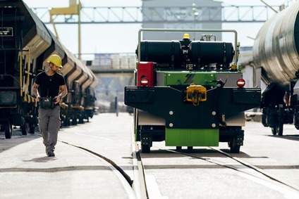 Worker in yellow helmet with remote control beside green shunting unit between freight wagons.
