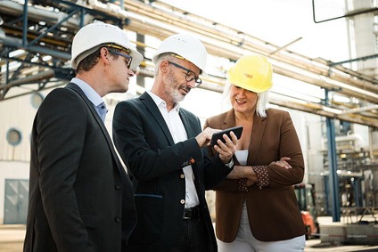 Drei Personen mit Schutzhelmen betrachten ein Smartphone auf einem Industriegelände.