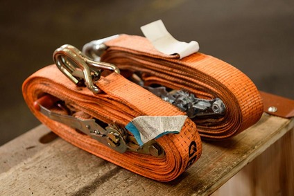 Aufgerollte orange Zurrgurte mit Metallratschen auf einer Holzoberfläche.