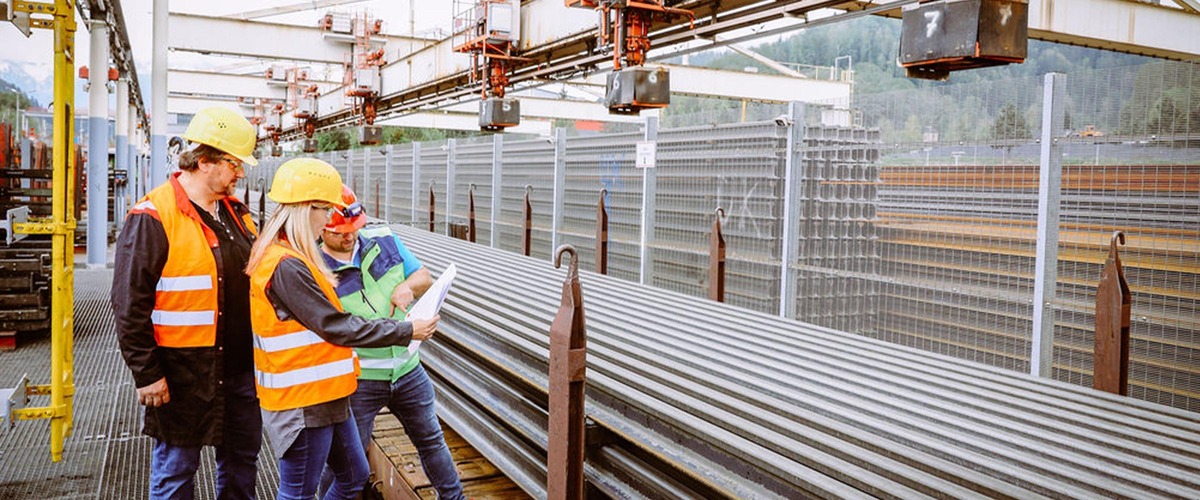 Three workers wearing safety helmets inspecting steel profiles in a production facility