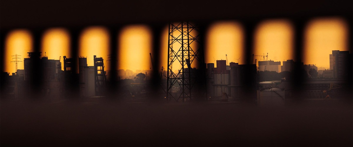 Industriegebiet bei Sonnenuntergang, Silhouetten von Fabriken und Strommasten durch Zaun im Vordergrund