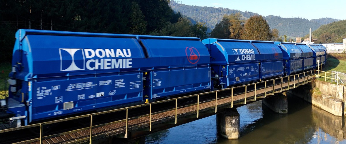 Blauer Donau Chemie Güterzug fährt über eine Brücke durch eine bewaldete Landschaft.