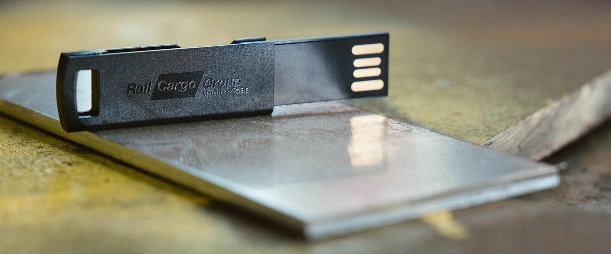 Schwarzer USB-Stick mit Rail Cargo Group Logo auf einer Metalloberfläche