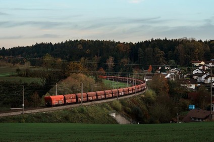 Güterzug mit roten Wagen fährt durch eine ländliche Landschaft und passiert Felder und Häuser in der Dämmerung