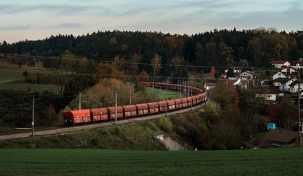 Langer Güterzug in einer Kurve nahe eines Dorfes, umgeben von herbstlichen Bäumen und Wiesen