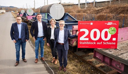 Vier Vertreter vor Güterwagen mit Stahlblöcken und Schild „200. Stahlblock auf Schiene“