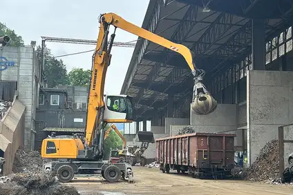 Bagger mit Greifer lädt Metallschrott in einen offenen Güterwagen in einer Recyclinganlage