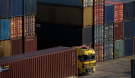 Gelber Lkw fährt entlang gestapelter Container in einem Containerterminal.
