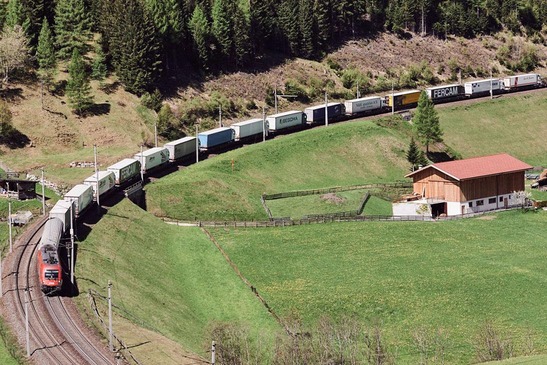 ROLA-Zug transportiert Lkw auf Bahnstrecke durch alpine Landschaft mit Wiesen und Wald