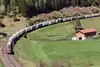 ROLA-Zug transportiert Lkw auf Bahnstrecke durch alpine Landschaft mit Wiesen und Wald